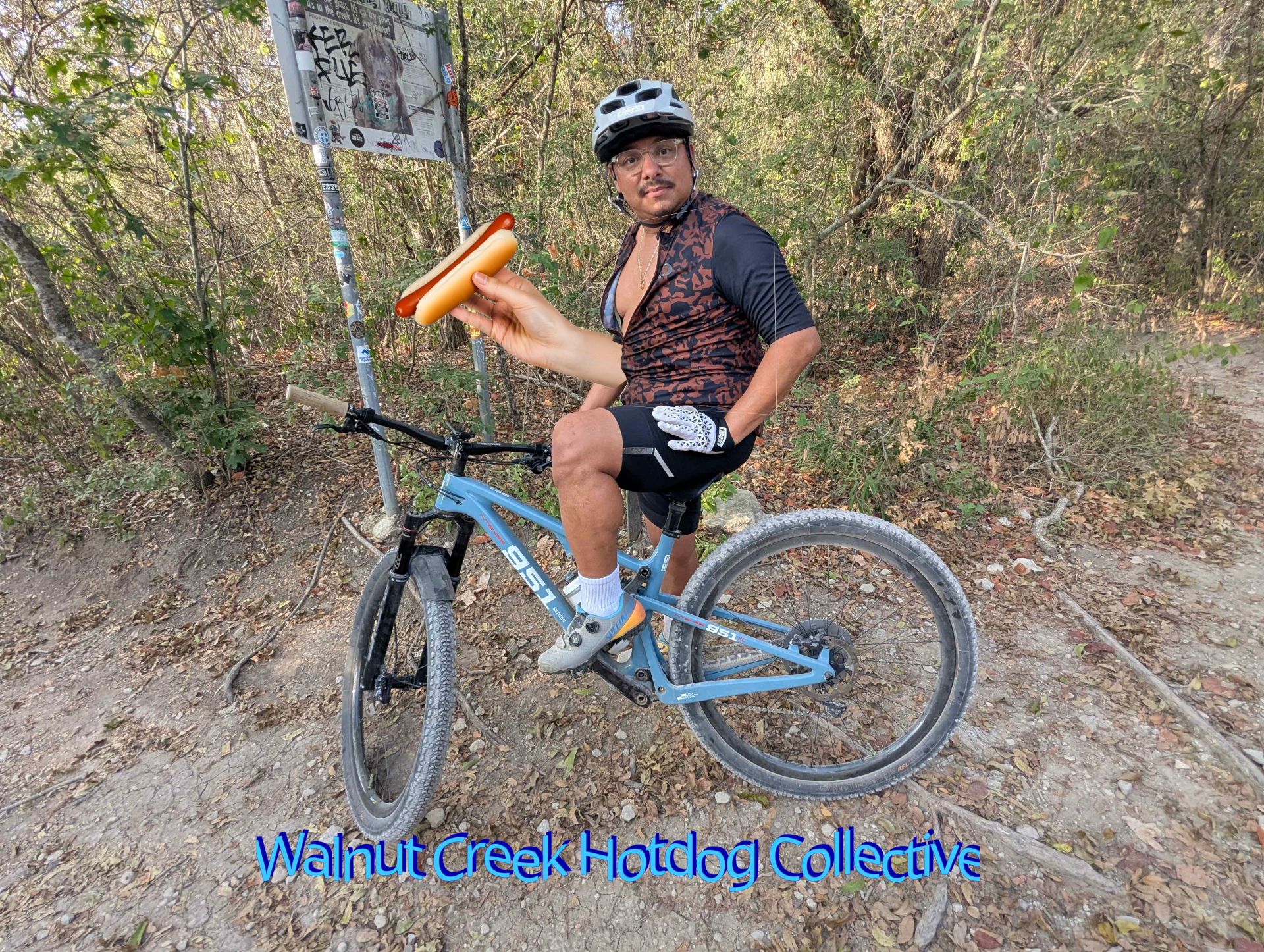 A man showing, a little skin, on a mountain bike, holding a hotdog with the words Walnut Creek Hotdog Collective Underneath him.